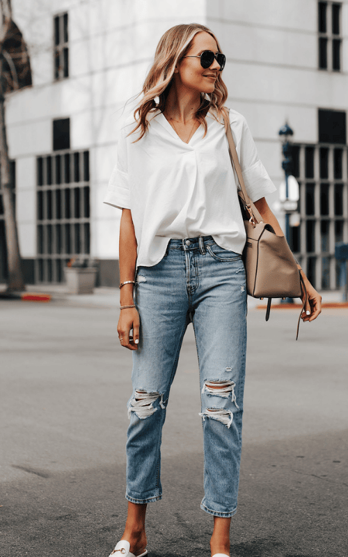 Woman wearing white loose top with ripped blue jeans, sunglasses, and handbag in casual street style